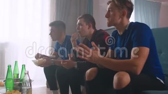 三个穿蓝色T恤的男人看足球赛朋友们坐在沙发上欢呼鼓掌视频的预览图