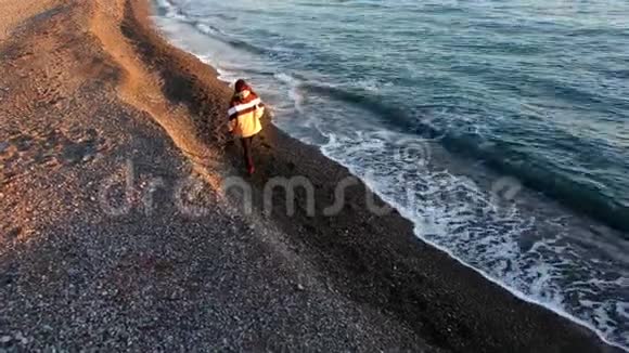 在夕阳的黄金时刻年轻女孩摄影师带着相机在海滩上散步视频的预览图