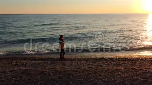 在夕阳的黄金时刻鸟瞰年轻女孩摄影师带着相机在海滩上散步视频的预览图