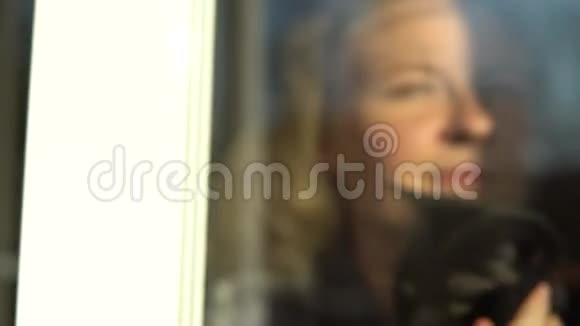 年轻的金发女人抱着一只猫微笑着看着窗外科罗纳病毒检疫期间呆在家里视频的预览图