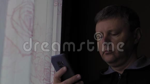 一个白人在一个黑暗的房间里靠窗户打电话视频的预览图