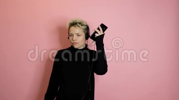 戴着智能手机戴着耳机在粉色背景下翩翩起舞的快乐无忧无虑的时尚女性视频的预览图