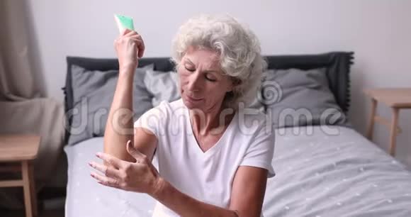 微笑的老年妇女在坐在床上的肘部涂抹奶油视频的预览图