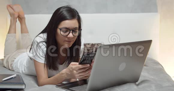 年轻女性在家工作时躺在床上使用智能手机和笔记本电脑视频的预览图