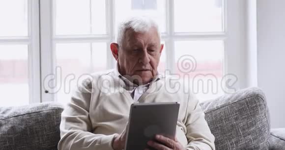 微笑老人用数码平板电脑在沙发上放松视频的预览图
