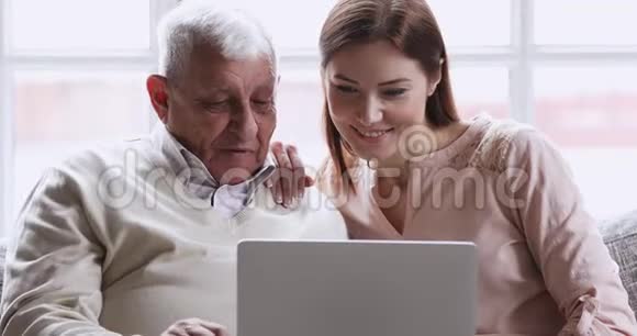 微笑着年轻的成年孙女用笔记本电脑教老爷爷视频的预览图