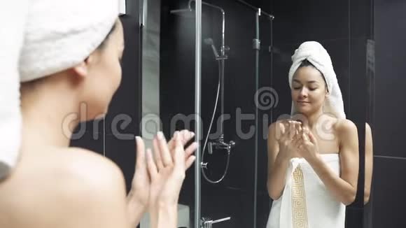 快乐的年轻女人头上拿着毛巾在浴室里涂手霜视频的预览图