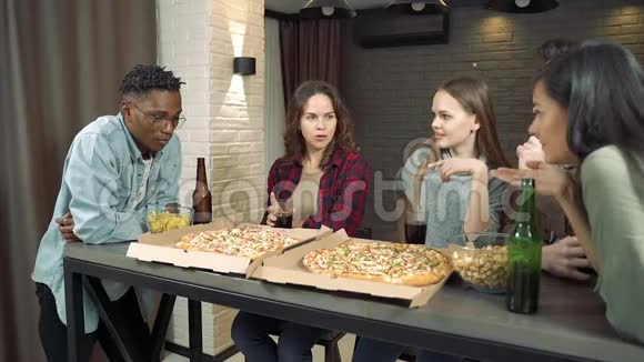 快乐的多民族朋友在家的客厅里喝啤酒吃披萨一起玩视频的预览图