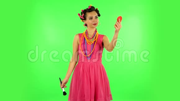 头上有卷发的女孩穿着一件粉红色的连衣裙看着红色的镜子用大刷子把鼻子涂成粉末绿色屏幕视频的预览图