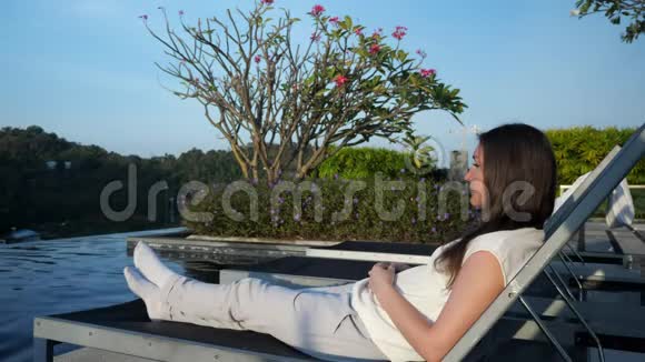 年轻女子躺在游泳池的折叠椅上坐在盛开的树旁视频的预览图
