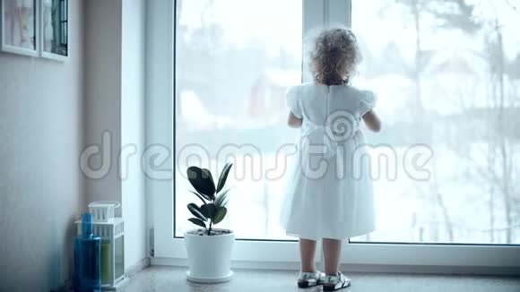 卷发的金发女婴穿着白色连衣裙看着窗外飘落的雪视频的预览图