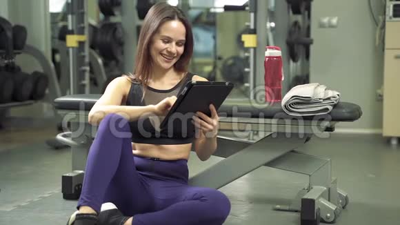 年轻的微笑女教练在健身房使用平板电脑制作在线培训项目视频的预览图