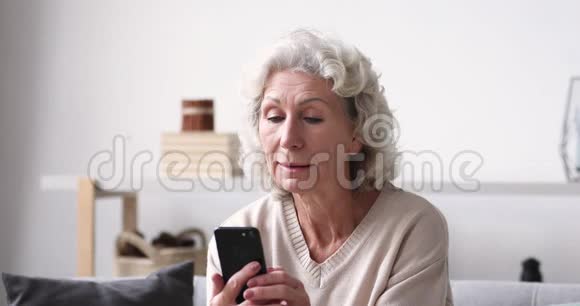 坐在沙发上用智能手机微笑着年长的祖母视频的预览图