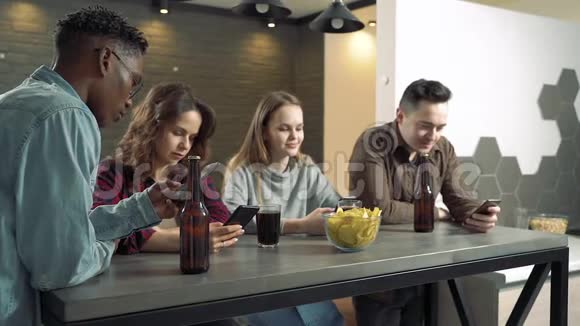 多民族的朋友群体积极使用手机4个人喝啤酒吃零食完全专注于他们的智能手机视频的预览图