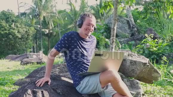 一个男人坐在岩石上一边看笔记本电脑上的视频一边用耳机听视频的预览图