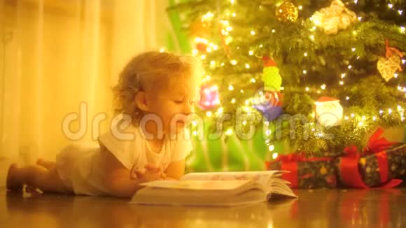 快乐的金发女婴在圣诞树旁的童话书中看照片视频的预览图