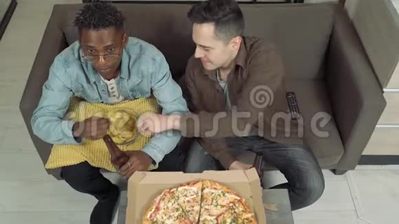 两个朋友在电视上看体育比赛男人在家坐在沙发上喝啤酒吃披萨和零食视频的预览图