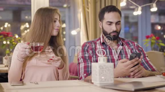 可爱的高加索女孩用智能手机看男朋友和大笑的肖像中东男人藏着电话视频的预览图
