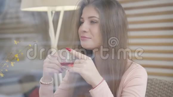 透过窗户拍摄一个积极的年轻白种人女人坐在咖啡馆里喝着茶微笑着Brunette视频的预览图