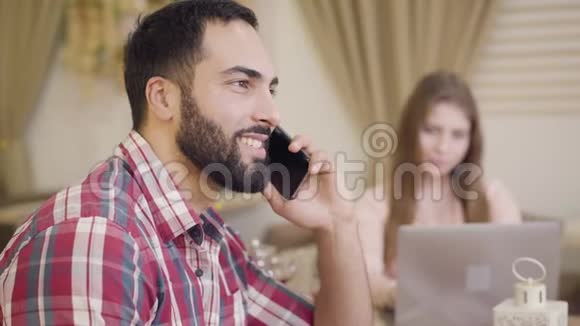 侧视微笑自信的中东商人在电话中交谈模糊的年轻白种人女人在工作视频的预览图