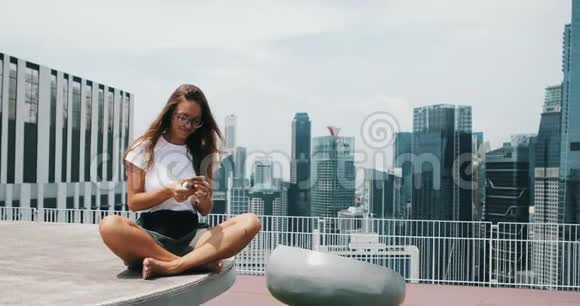 一个漂亮的年轻女孩穿着一件白色T恤手机背景是高楼大厦和城市摩天大楼视频的预览图