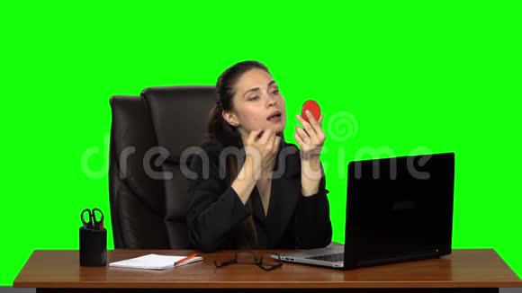 商务女性坐在皮椅上的办公桌前用红色的镜子描绘她的嘴唇绿色屏幕慢动作视频的预览图