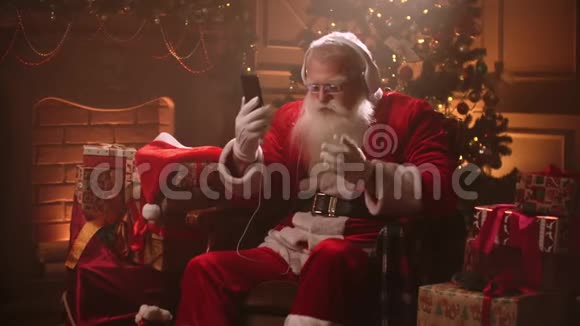戴着耳机和手里拿着手机跳舞的圣诞老人在线听音乐充满活力的歌曲视频的预览图
