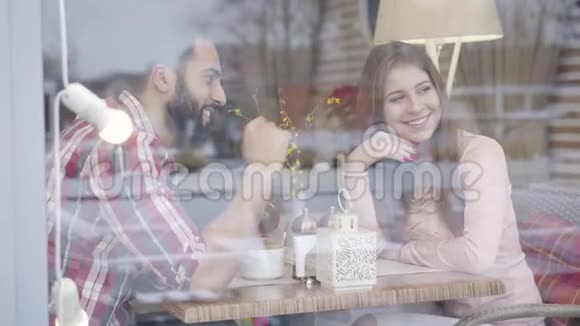 透过窗户拍摄一对年轻的多民族夫妇在咖啡馆里坐在桌子旁交谈微笑视频的预览图