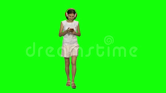 年轻的女人正平静地戴着红色的大耳机行走在绿色屏幕上的手机上选择音乐前景视频的预览图