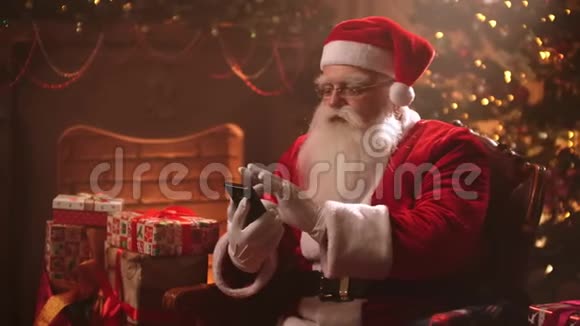 圣诞老人坐在圣诞树背景的椅子上用智能手机查看他的网上银行视频的预览图