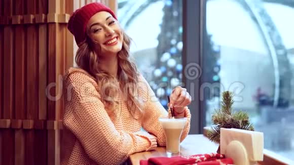 美丽的白种人金发女郎戴着红色针织帽子坐在自助餐厅的窗户后面喝着一杯温暖的拿铁视频的预览图