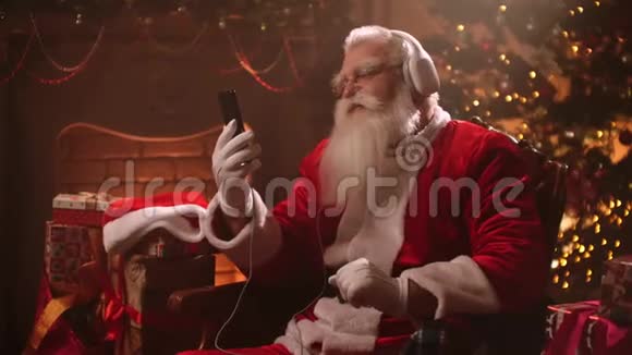 圣诞前夜留着白胡子的老人带着耳机和圣诞老人服装听音乐圣诞老人视频的预览图