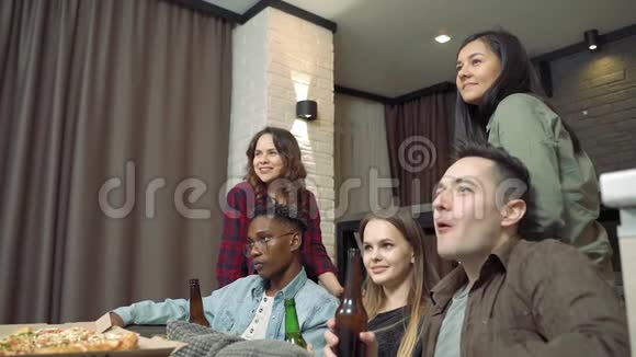 一群多民族的朋友在家的客厅里一起喝啤酒一起看电视视频的预览图