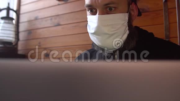 戴着医用防护面具的大胡子男子用笔记本电脑从网上阅读有关covid19的新闻男性关心他的h视频的预览图
