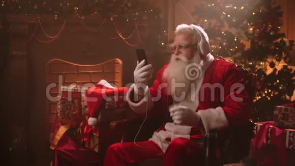 戴着耳机和手里拿着手机跳舞的圣诞老人在线听音乐充满活力的歌曲视频的预览图