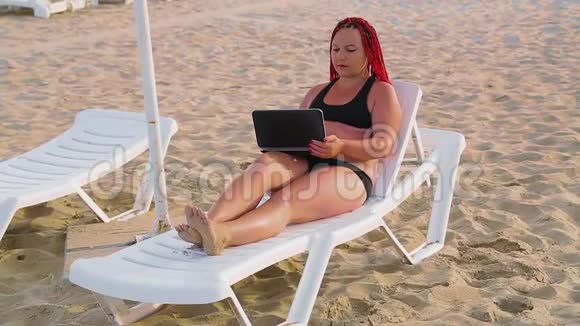 穿着泳衣在沙滩上的年轻女子她坐在躺椅上手提电脑视频的预览图