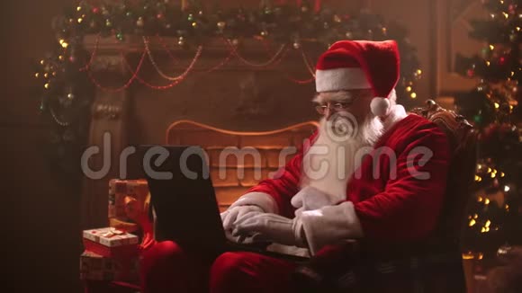 一位身穿圣诞老人服装的老人正在圣诞树背景的笔记本电脑前工作视频的预览图