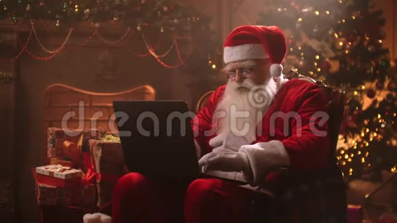 有着白胡子的圣诞老人坐在椅子上在笔记本电脑上工作阅读孩子们的电子邮件视频的预览图