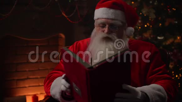 穿着圣诞老人服装的老人坐在大气的房间里读着一本红色封面的神奇闪亮的书视频的预览图