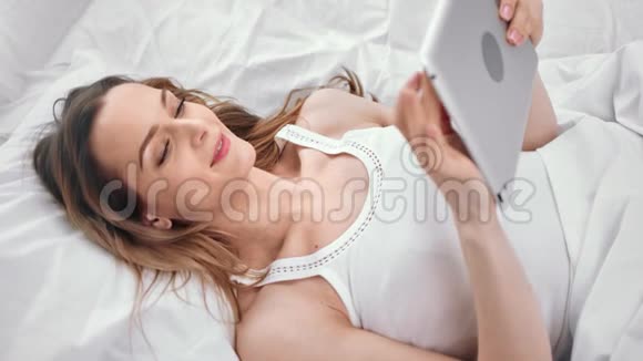 微笑的年轻女孩看着躺在床上的平板电脑屏幕近距离拍摄4k红色相机视频的预览图