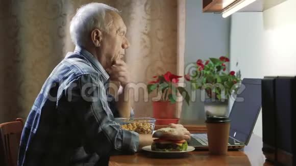一位老人在家放松晚上用笔记本电脑看新闻或电视节目吃爆米花和汉堡视频的预览图