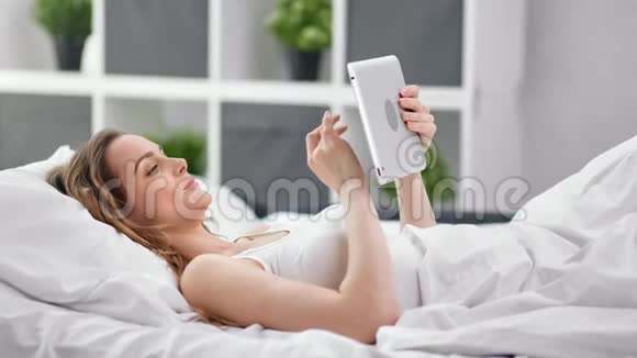 愉快的年轻女子躺在床上聊天使用平板电脑享受周末4K红色相机拍摄视频的预览图