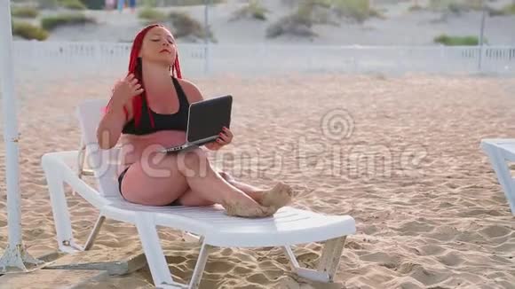 穿着泳衣在海滩上的一个带着笔记本电脑的躺椅上的年轻女人红头发视频的预览图