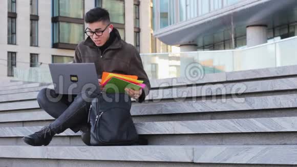 年轻快乐的亚洲男学生拿着笔记本电脑坐在台阶上视频的预览图