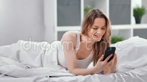 微笑的年轻女子躺在床上用智能手机轻松聊天4K红色摄像机拍摄视频的预览图