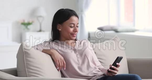 在沙发上用智能手机微笑轻松的非洲年轻女性视频的预览图