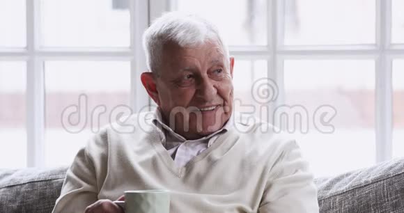 快乐的老人白发苍苍的男人在沙发上喝茶放松视频的预览图