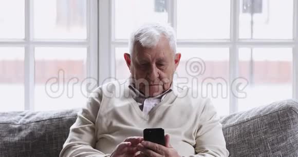 微笑的80年代爷爷坐在沙发上使用手机应用程序视频的预览图