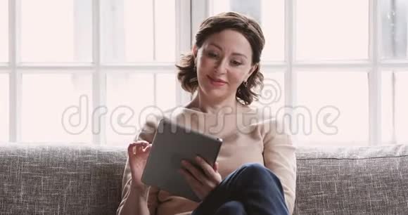 使用数码平板电脑放松的中年妇女坐在沙发上视频的预览图
