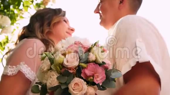新娘和新郎在茂密的白玫瑰灌木丛附近微笑和亲吻新婚夫妇拥抱在一起彼此相爱视频的预览图